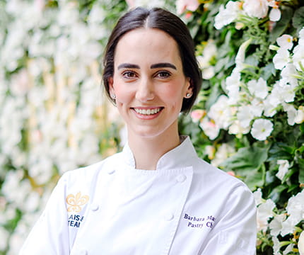 The Little Nell Pastry Chef, Barbara Marcos, in her chef coat in front of a flower wall.